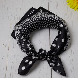 May include: A black and white bandana with a paisley and polka dot pattern is knotted. The square scarf is folded and tied, showcasing the contrasting designs. The accessory is made of a silky material.