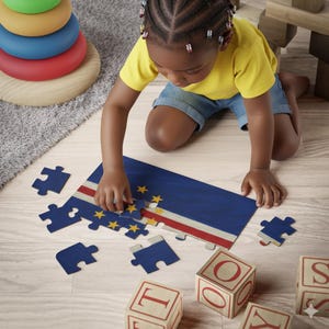 Puede incluir: Un niño está armando un rompecabezas que representa la bandera de Cabo Verde, con azul, blanco, rojo y estrellas amarillas. Bloques de letras de madera están dispersos. El niño lleva una camiseta amarilla y pantalones cortos de mezclilla.
