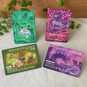 Peut inclure: Quatre cartes de jeu rectangulaires avec des illustrations colorées. En haut à gauche : carte verte TENTACLE avec une créature dans des tentacules. En haut à droite : carte violette SHADOW CLONE avec une silhouette encapuchonnée. En bas : cartes BRUSH et BARON PIT.