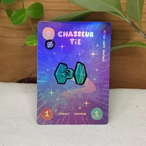 Può includere: Una carta olografica con un caccia TIE verde acqua su uno sfondo spaziale viola e blu. La carta presenta il testo "CHASSEUR TIE" e altro testo francese, con il numero 1 in un cerchio negli angoli inferiori.