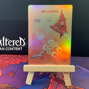 May include: A holographic trading card with the text "SOL and HALUA" and an illustration of a person in a red sailplane. The card also depicts a bird and stylized clouds. The card is displayed on a small wooden easel.