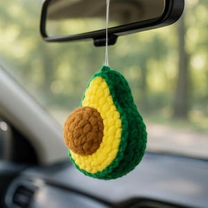 Könnte beinhalten: Ein gehäkelter Avocado-Autoanhänger, der am Rückspiegel hängt. Der Anhänger hat eine grüne Außenseite, eine gelbe Innenseite und einen braunen Kern. Der Anhänger wird von einer weißen Schnur gehalten und verleiht dem Innenraum des Autos einen handgefertigten Charme.