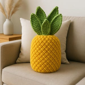 May include: A handmade, yellow and green crochet pineapple pillow. The pillow has a textured, yellow body and green leaves. The pillow is sitting on a beige couch with a white pillow behind it.