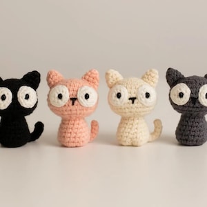 Może przedstawiać: Cztery ręcznie robione figurki kotów na szydełku w kolorze czarnym, różowym, kremowym i szarym. Każdy kot ma duże białe oczy, mały czarny nos i zakręcony ogon. Koty ustawione są w rzędzie na gładkim tle.