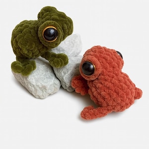 May include: Two handmade crochet frog plushies. One is green, the other is orange-red. Both have large black eyes with gold rims and are posed on grey rocks. The frogs have textured bodies and are made of soft yarn.
