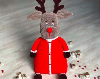 May include: A handmade plush reindeer toy with a red Santa-style coat, brown antlers, and a red nose. The reindeer is wearing a red coat with white trim and buttons. The toy is surrounded by small gift boxes and red confetti.
