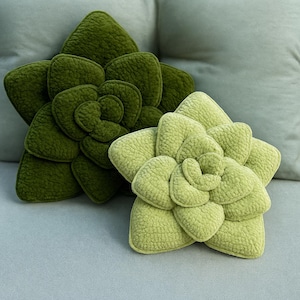 Puede incluir: Dos cojines verdes con forma de suculenta. Uno es de un tono verde más oscuro, el otro de un verde más claro, casi lima. Los cojines tienen un aspecto texturizado y aterciopelado y están colocados sobre una superficie gris claro, lo que sugiere un elemento decorativo para el hogar.