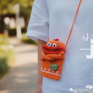 Può includere: Borsa a tracolla all'uncinetto arancione con un design a forma di anatra dei cartoni animati. La borsa presenta un becco rosso, occhi bianchi e una piccola tasca verde. La tracolla arancione è indossata su una camicia bianca.