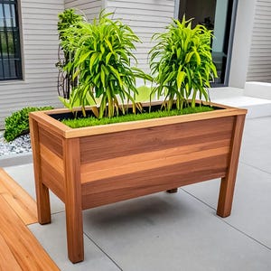 May include: A rectangular wooden planter box with a rich, natural wood grain finish. The planter is filled with green plants and sits on four sturdy legs. The design is modern and suitable for outdoor use.