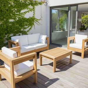 Puede incluir: Juego de muebles de exterior de madera con cojines blancos. El conjunto incluye un sofá de dos plazas, dos sillones y una mesa de centro rectangular. Los muebles están sobre una terraza de madera, con una pared cubierta de hiedra verde al fondo.