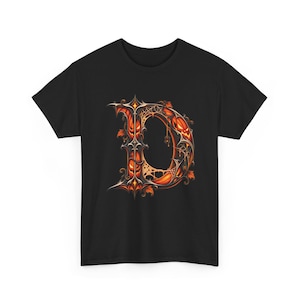 Könnte beinhalten: Ein schwarzes T-Shirt mit einem großen, verzierten Buchstaben D als Grafik. Der Buchstabe ist mit leuchtend orangefarbenen Kürbisgesichtern, komplexen, dunkelmetallischen Ornamenten, Spinnennetzen und kleinen Herbstblättern gestaltet.