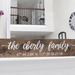 May include: A wooden sign with the text "the cberly family" in white script. Below the text are the coordinates 47° 44' 2.64" N 117° 28' 59.21" W. A clear glass bottle with purple flowers is to the left.
