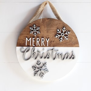 K&ouml;nnte beinhalten: Ein rundes Holzschild mit einer braunen oberen H&auml;lfte und einer wei&szlig;en unteren H&auml;lfte. Das Schild tr&auml;gt die Aufschrift "Frohe Weihnachten" in wei&szlig;en Buchstaben und hat drei wei&szlig;e Schneeflocken-Ausschnitte. Das Schild h&auml;ngt an einem Juteschnur.