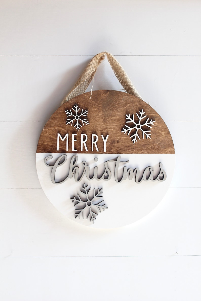 K&ouml;nnte beinhalten: Ein rundes Holzschild mit einer braunen oberen H&auml;lfte und einer wei&szlig;en unteren H&auml;lfte. Das Schild tr&auml;gt die Aufschrift "Frohe Weihnachten" in wei&szlig;en Buchstaben und hat drei wei&szlig;e Schneeflocken-Ausschnitte. Das Schild h&auml;ngt an einem Juteschnur.