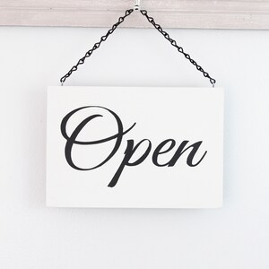 May include: A white sign with black cursive lettering that reads "Open" hanging from a black chain on a white wall.