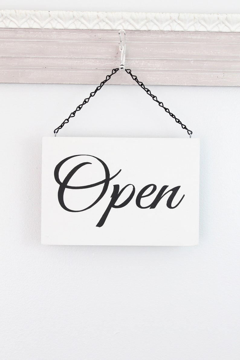 May include: A white sign with black cursive lettering that reads "Open" hanging from a black chain on a white wall.