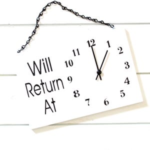 May include: White sign with black text that reads "Will Return At" and a clock face with black hands. The sign is hanging from a black chain.