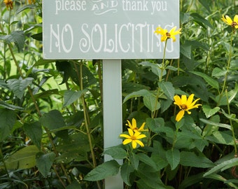 May include: A white sign with the text "please and thank you no soliciting" in black lettering, mounted on a wooden stake, surrounded by yellow flowers and green foliage.