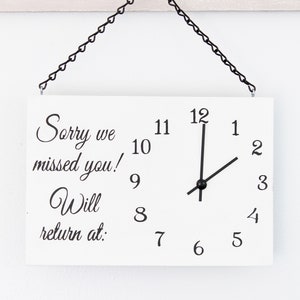 May include: White wooden sign with black text that reads "Sorry we missed you! Will return at:" and a clock face with black hands set to 12:00. The sign is hanging from a black chain.