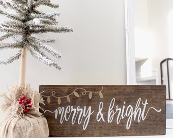May include: A brown wooden sign with the text "merry & bright" in white paint. The sign has a gold painted string of lights above the text. The sign is sitting on a white mantel with a small, snowy artificial Christmas tree to the left.