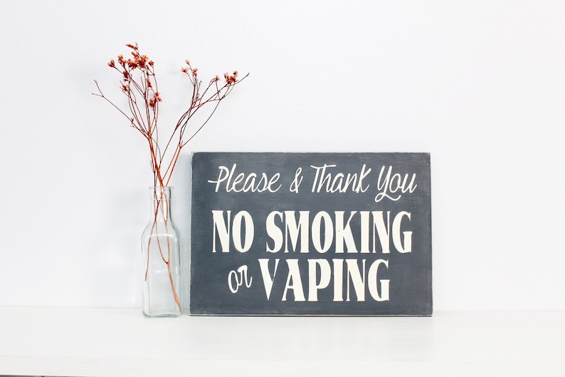 Może przedstawiać: Szary drewniany znak z białym napisem "Please & Thank You NO SMOKING or VAPING". Znak stoi na białym tle, obok przezroczystego szklanego wazonu z suszonymi kwiatami.