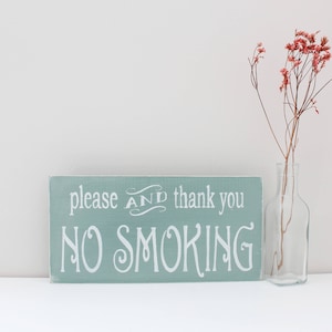 Puede incluir: Un letrero de madera azul claro con texto blanco que dice "please AND thank you NO SMOKING". El letrero est&aacute; sobre una superficie blanca junto a un jarr&oacute;n de vidrio transparente con flores rosadas secas.