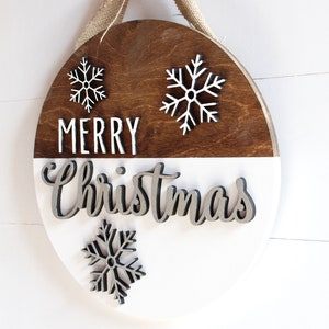 K&ouml;nnte beinhalten: Ein rundes Holzschild mit wei&szlig;er und brauner Lackierung. Das Schild tr&auml;gt die Aufschrift "Merry Christmas" in wei&szlig;en und silbernen Buchstaben. Auf dem Schild befinden sich drei wei&szlig;e Schneeflocken-Ausschnitte.