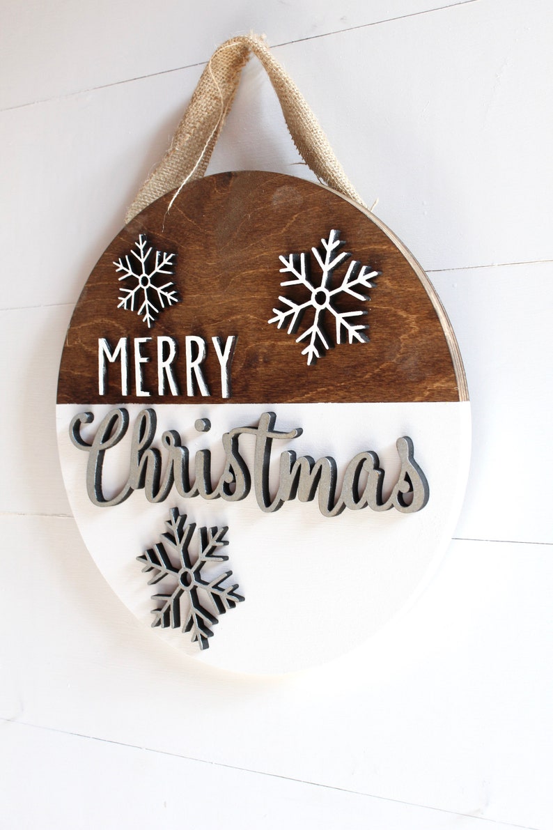 K&ouml;nnte beinhalten: Ein rundes Holzschild mit wei&szlig;er und brauner Lackierung. Das Schild tr&auml;gt die Aufschrift "Merry Christmas" in wei&szlig;en und silbernen Buchstaben. Auf dem Schild befinden sich drei wei&szlig;e Schneeflocken-Ausschnitte.