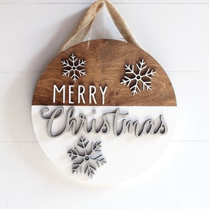K&ouml;nnte beinhalten: Ein rundes Holzschild mit wei&szlig;er und brauner Bemalung. Das Schild sagt "Merry Christmas" mit Schneeflocken oben und unten. Das Schild hat einen Jute-Aufh&auml;nger.