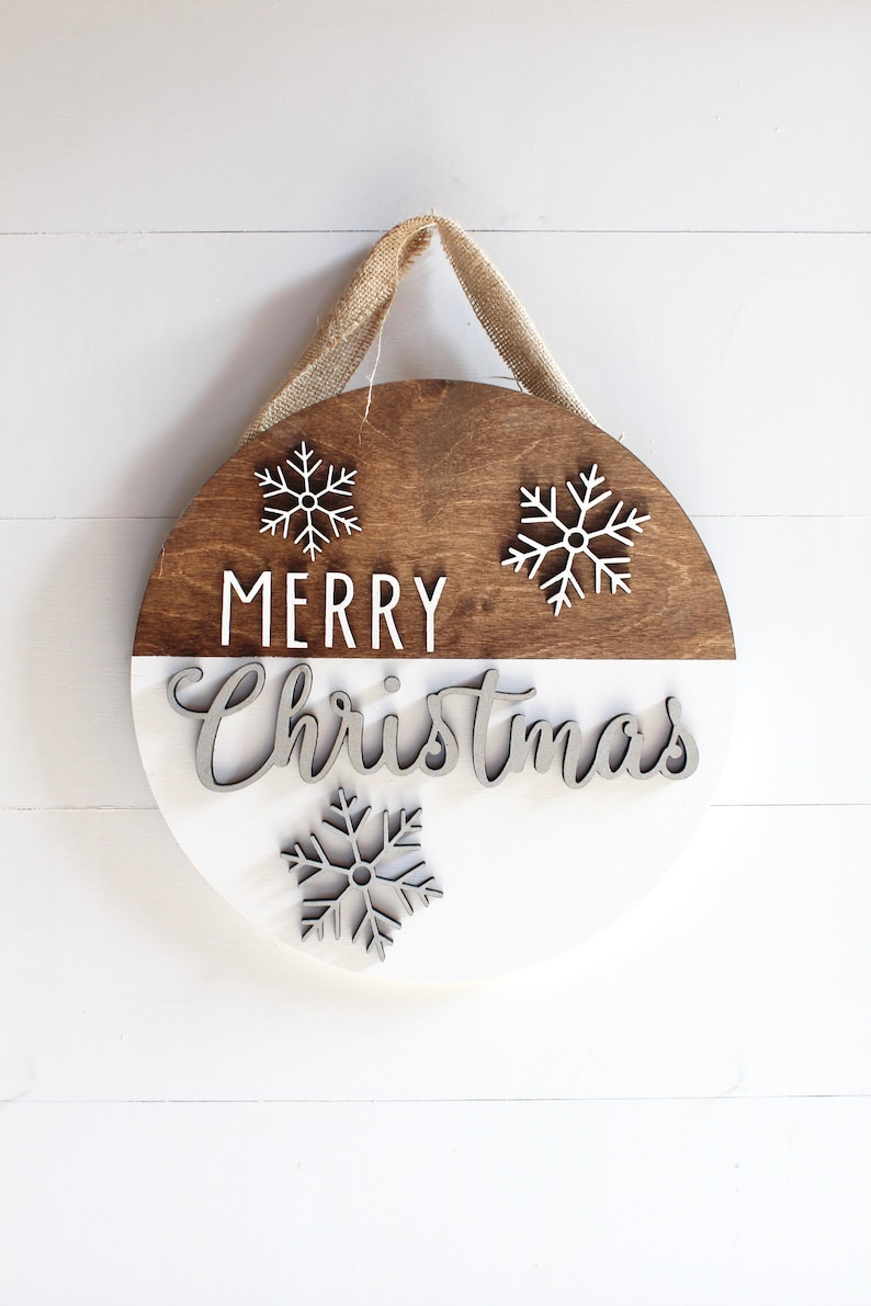 K&ouml;nnte beinhalten: Ein rundes Holzschild mit wei&szlig;er und brauner Bemalung. Das Schild sagt "Merry Christmas" mit Schneeflocken oben und unten. Das Schild hat einen Jute-Aufh&auml;nger.