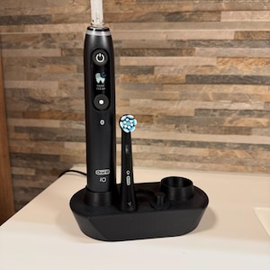 Puede incluir: Cepillo de dientes eléctrico Oral-B iO negro con base de carga y dos cabezales. El cepillo de dientes tiene una pantalla digital y un botón de encendido. El texto "DAILY CLEAN" es visible en la pantalla. La base tiene un soporte para los cabezales.