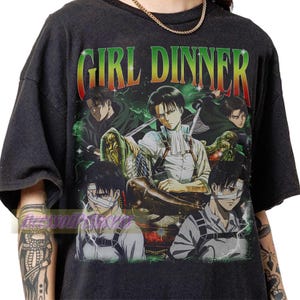 Op de afbeelding: Een zwart t-shirt met een grafische print met de tekst "GIRL DINNER" in een rood, geel en groen verlooplettertype. Het ontwerp toont meerdere illustraties van een donkerharig anime-personage in verschillende houdingen met een wit hemd.