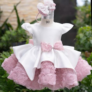 May include: A white dress with short sleeves and a pink sequined bow at the waist. The skirt features layered pink fabric rosettes. A matching pink lace headband with a bow sits on a mannequin torso marked with the number 90 on the neck.