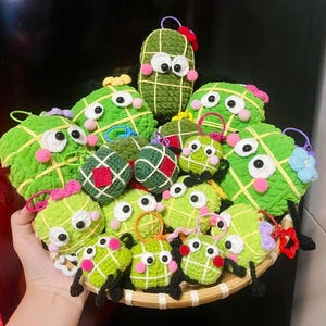 May include: A collection of handmade green crochet ornaments with cartoon faces, including large and small sizes. The ornaments have white eyes, pink cheeks, and are arranged in a woven basket. Some have colorful accents.