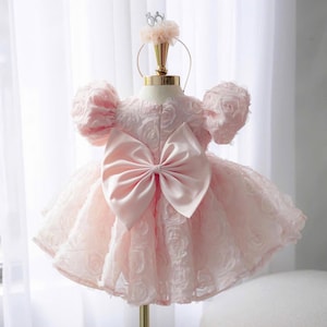 May include: A pale pink dress featuring a textured rose-patterned fabric, short puff sleeves, and a large satin bow centered on the back. The garment is displayed on a mannequin stand topped with a matching pink floral headband accessory.