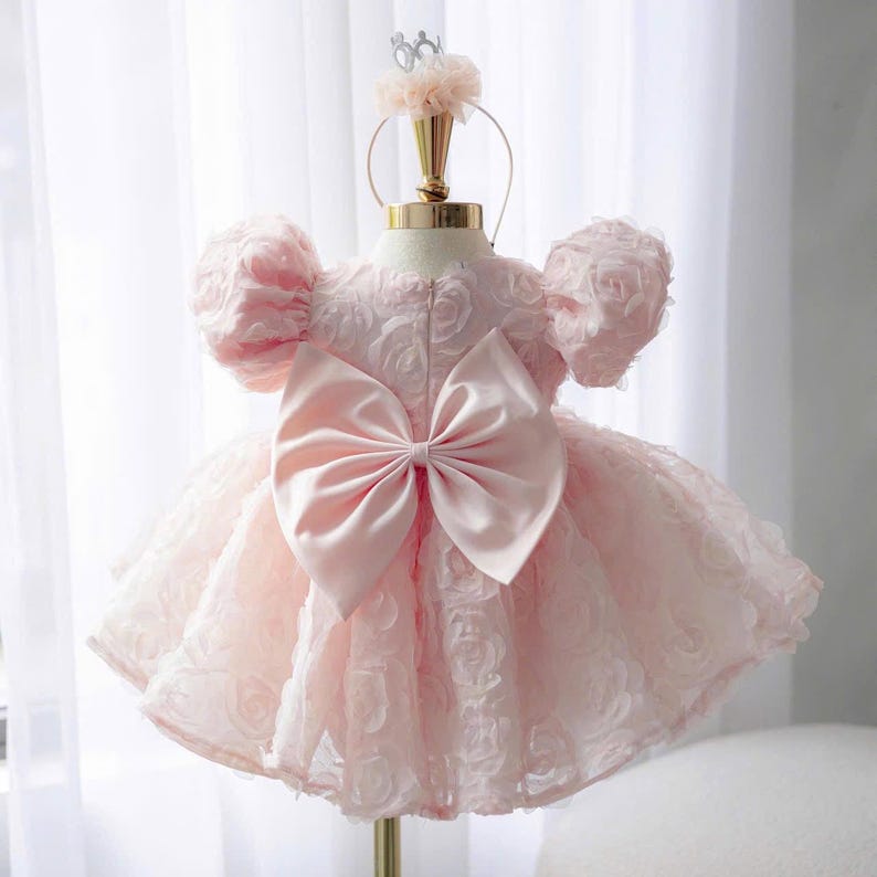 May include: A pale pink dress featuring a textured rose-patterned fabric, short puff sleeves, and a large satin bow centered on the back. The garment is displayed on a mannequin stand topped with a matching pink floral headband accessory.