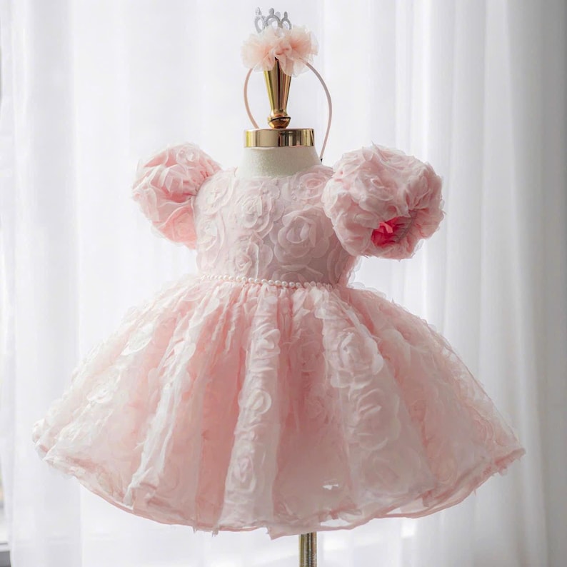 May include: A pale pink dress featuring short puff sleeves and a bodice covered in layered fabric rosettes. The waistline is accented with a delicate row of small white pearls. A matching headband with a pink floral embellishment is positioned above the dress on a display stand.