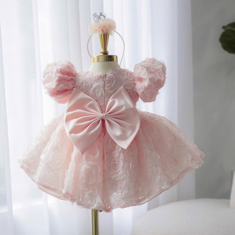 May include: A pale pink dress featuring a textured rose pattern, short puff sleeves, and a large satin bow centered on the back. The garment is displayed on a mannequin stand topped with a matching pink floral headpiece accessory.