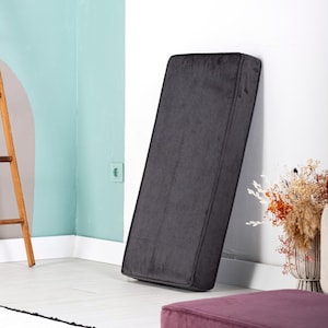 May include: A dark gray rectangular cushion with rounded corners, leaning against a white wall. The cushion has a soft, velvety texture. The room has a modern design with a light blue wall and a wooden ladder.