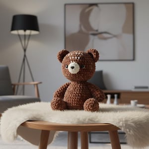 May include: A brown crochet teddy bear with a white muzzle and head markings. The bear sits on a small wooden table covered with a white faux fur rug. The background features a lamp, chair, and artwork, creating a cozy, domestic scene.