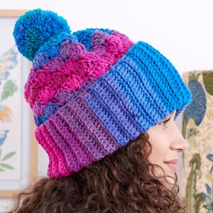 Puede incluir: Un gorro de ganchillo hecho a mano y colorido con un pompón. El gorro presenta un degradado de colores, incluyendo azul, rosa y morado. El borde es azul liso y el pompón es azul vibrante. El gorro está diseñado para la calidez y el estilo.