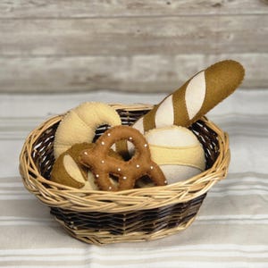 Puede incluir: Una cesta de mimbre llena de productos de pan de fieltro. La cesta contiene un cruasán, un pretzel, pan redondo y una baguette. Los productos de pan son en tonos marrones y beige, con detalles blancos. La cesta está sobre una superficie rayada.