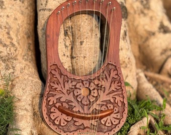 May include: A handcrafted wooden lyre with intricate floral carvings. The instrument features a curved soundbox and a series of strings. The lyre is a reddish-brown color and rests against a tree trunk.