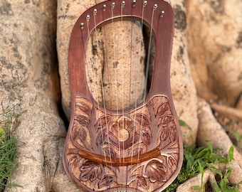 May include: A handcrafted wooden lyre with a carved floral design. The instrument has a dark brown finish, with strings stretched across the soundboard. The lyre is set against a natural background of tree roots and foliage.