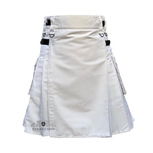 Puede incluir: Un kilt blanco con detalles negros, con pliegues y correas ajustables. El kilt tiene bolsillos y anillas en D para accesorios. El texto "MEN KILT SHOP" es visible en la parte inferior.