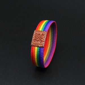 Peut inclure: Bracelet en tissu rayé arc-en-ciel avec un patch en cuir marron. Le patch présente un logo avec les lettres "RQ" et un motif en croix. Les couleurs du bracelet comprennent le rouge, l'orange, le jaune, le vert, le bleu et le violet.