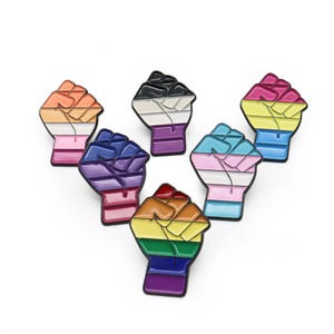 Könnte beinhalten: Sechs Emaille-Anstecknadeln in Form einer geballten Faust, jede mit einem anderen LGBTQ+-Pride-Flaggen-Design. Die Anstecknadeln haben eine schwarze Umrandung und sind fächerförmig auf weißem Hintergrund angeordnet.