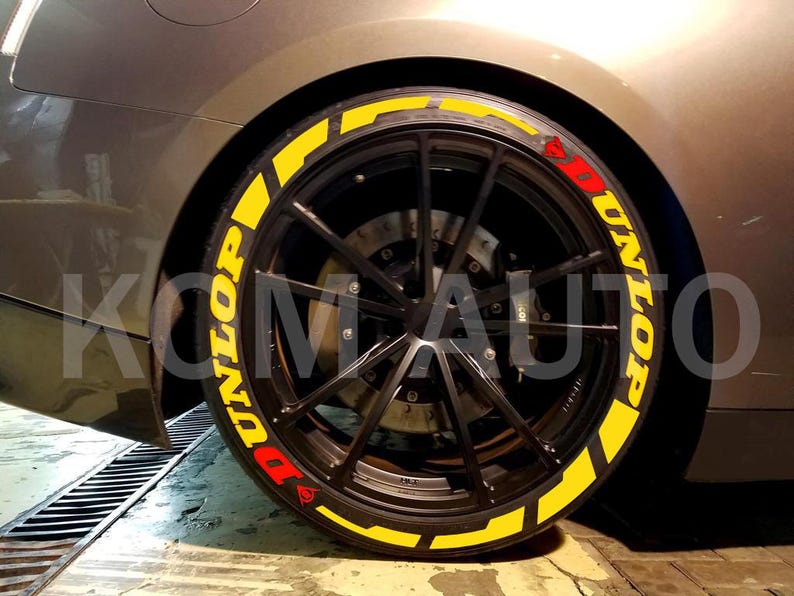 May include: Close-up of a car wheel with a black rim and a tire featuring yellow and red Dunlop branding. The tire has a black sidewall with yellow lettering and accents. The car's body is gray.