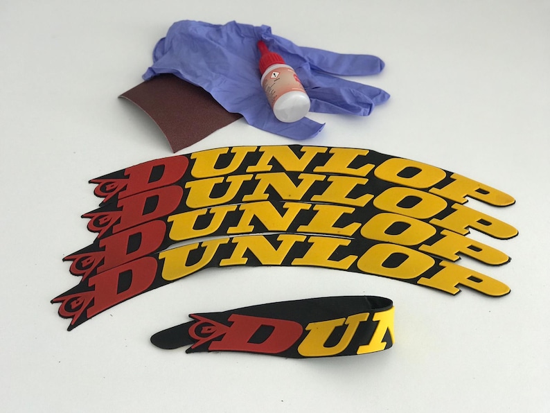 May include: A collection of Dunlop motorcycle tire decals in red, yellow, and black. Includes a pair of blue gloves, a small bottle of adhesive, and a piece of sandpaper. The decals are curved and feature the Dunlop logo.