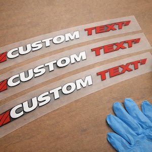 May include: Four clear decals with the words "CUSTOM TEXT" in white and red lettering. A blue bottle with a black cap and a pair of blue gloves are also in the image. The decals are for customization.